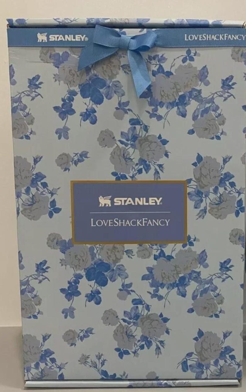 May include: A box with a floral pattern in light blue and white, tied with a blue bow. The box displays the "STANLEY" and "LOVESHACKFANCY" logos. The floral design features white roses and blue leaves.