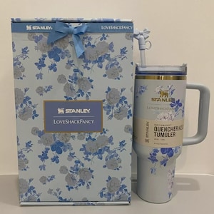 May include: A light blue Stanley Loveshackfancy Quencher H2.0 tumbler with a matching gift box. Both feature a floral pattern in blue and white. The tumbler has a handle and straw. The box is tied with a blue ribbon.