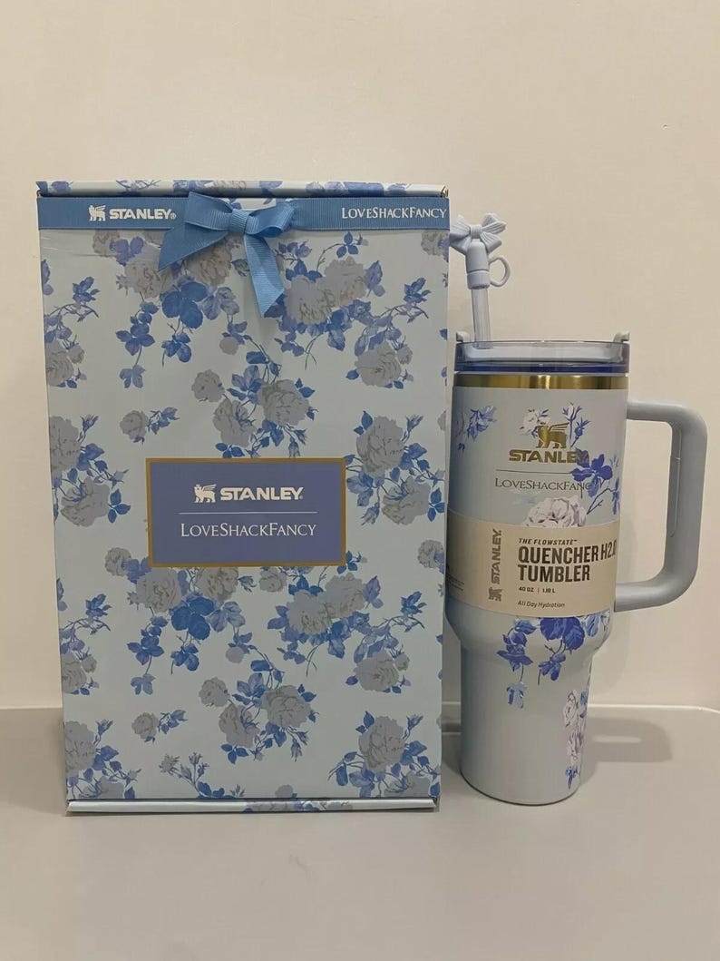 May include: A light blue Stanley Loveshackfancy Quencher H2.0 tumbler with a matching gift box. Both feature a floral pattern in blue and white. The tumbler has a handle and straw. The box is tied with a blue ribbon.