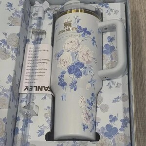May include: A white Stanley travel mug with a handle, decorated with blue and pink floral patterns. It comes in a box with a matching floral design, a straw, and a cleaning brush. The text "STANLEY LoveShackFancy" is visible.