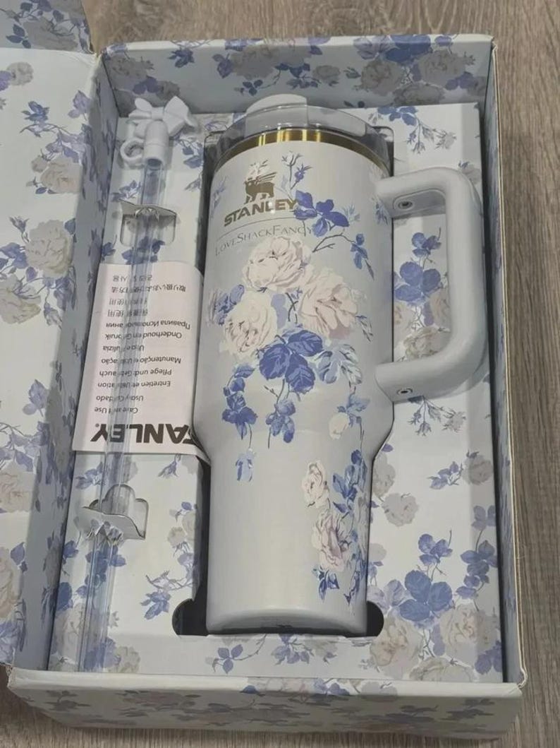 May include: A white Stanley travel mug with a handle, decorated with blue and pink floral patterns. It comes in a box with a matching floral design, a straw, and a cleaning brush. The text "STANLEY LoveShackFancy" is visible.
