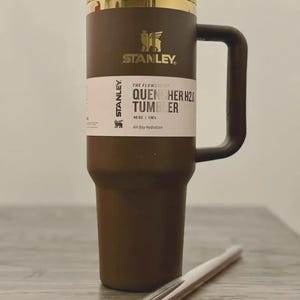 Puede incluir: Un vaso Stanley Quencher H2.0 marr&oacute;n con tapa y asa doradas. El vaso tiene una etiqueta blanca con el texto "THE FLOWSTATE QUENCHER H2O TUMBLER" y "40 OZ | 1.18 L". Una pajita transparente descansa frente al vaso.