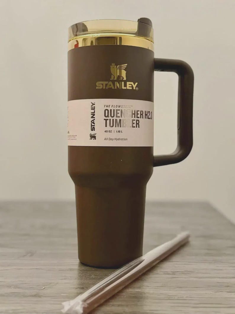 Puede incluir: Un vaso Stanley Quencher H2.0 marr&oacute;n con tapa y asa doradas. El vaso tiene una etiqueta blanca con el texto "THE FLOWSTATE QUENCHER H2O TUMBLER" y "40 OZ | 1.18 L". Una pajita transparente descansa frente al vaso.
