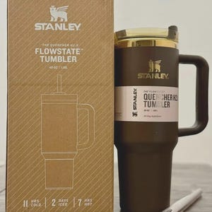 Puede incluir: Un vaso Stanley Flowstate marr&oacute;n con tapa y asa doradas, junto a su caja de cart&oacute;n. La caja y el vaso presentan el logotipo de Stanley y detalles del producto, incluyendo "40 oz | 1.18L". El vaso est&aacute; dise&ntilde;ado para la hidrataci&oacute;n durante todo el d&iacute;a.