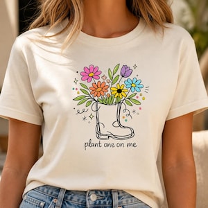 May include: A cream-colored t-shirt featuring a graphic of a black line-art rain boot filled with colorful flowers in pink, orange, yellow, purple, and blue. The text "plant one on me" is printed in black below the boot, surrounded by small dots and stars.
