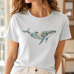 May include: A light blue short-sleeve t-shirt featuring a graphic of a whale on the chest. The whale illustration is composed of various patterns and textures in shades of blue, green, orange, and white, depicting coral and botanical elements.