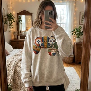Könnte beinhalten: Ein hellgraues Sweatshirt mit einem farbenfrohen, gemusterten Videospiel-Controller-Design. Der Controller hat rote, orange, blaue und grüne Akzente, mit einem Herz und einer Tasse Kaffee. Das Sweatshirt ist ein lässiger, bequemer Stil.