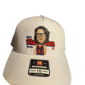 May include: White trucker hat featuring a cartoon image of Jon Fishman with the text "Jon Fishman" and "Phish". The hat has a curved brim and a mesh back. A black and white Richardson tag is on the front.