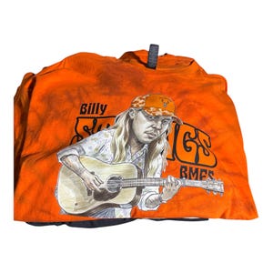 Può includere: Una t-shirt arancione tie-dye con un'illustrazione grafica di una persona dai capelli lunghi che suona una chitarra acustica. La persona indossa un berretto da baseball mimetico. Il testo "Billy Strings" e "BMFS" è stampato in nero.