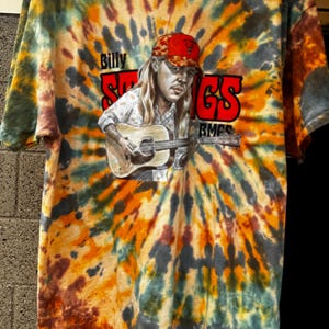 May include: A short-sleeve tie-dye t-shirt in shades of orange, yellow, green, and dark gray. The front features a graphic of a person with long hair wearing a red cap, playing an acoustic guitar, with the text Billy Strings BMFS.