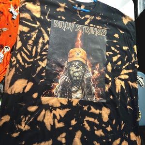 May include: A black short-sleeve t-shirt with a bleach-dyed spiral pattern. The center features a graphic print with the text "BILLY STRINGS" above an illustration of a figure wearing an orange cap, surrounded by lightning and chains.