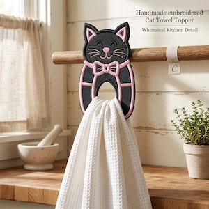 May include: A black cat-shaped towel topper with pink embroidered details, including ears, whiskers, and a bow tie, hanging on a wooden rod. A white waffle-weave towel is draped through the center opening of the cat-shaped holder.