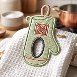 Puede incluir: Un paño de cocina blanco con tejido de gofre que presenta un parche bordado de una manopla de cocina en color verde salvia. El parche incluye un corazón rosa dentro de un cuadrado, un centro ovalado gris oscuro y un lazo color canela.