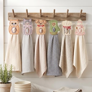 May include: Six decorative kitchen towels hanging on a wooden rack. Each towel features a unique embroidered topper shaped like an owl, cat, dog, oven mitt, chef hat, or apron. The towels vary in color and texture, including waffle weave and gingham.