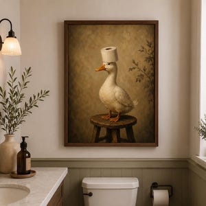 May include: A framed wall art print featuring a white duck with an orange beak standing on a small wooden stool. The duck has a roll of white toilet paper balanced on its head. The background is a muted, textured brown with faint leaf patterns.