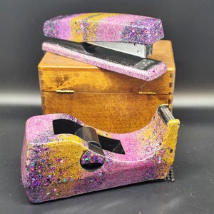 May include: A stapler and tape dispenser with a glittery pink, purple, and gold design. The stapler sits atop a small wooden box, while the tape dispenser rests below. The office supplies have a unique, decorative appearance.