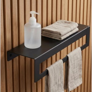 Könnte beinhalten: Ein schwarzes Metallregal mit integrierter Handtuchstange, montiert an einer Holzwand. Auf dem Regal steht ein klarer, mattierter Glas-Seifenspender, zusammen mit einem Stapel gefalteter beiger Handtücher. Zwei beige Handtücher hängen an der Handtuchstange.
