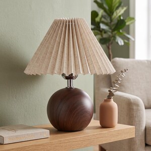 Peut inclure: Une lampe de table avec un abat-jour plissé beige et une base sphérique marron foncé. La lampe est posée sur une étagère en bois clair à côté d'un petit vase pêche et d'un livre intitulé "Scandinavian Design".