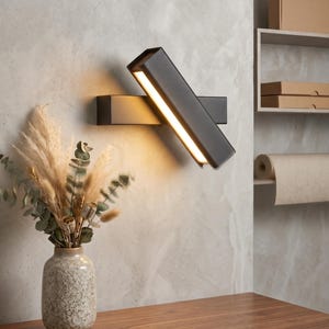 May include: Two contemporary, dark grey wall lights with integrated LED illumination. The rectangular lights are angled, emitting a warm glow. A decorative vase with dried flowers sits on a wooden table. The background is a textured, light grey wall.