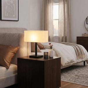 Pode incluir: Um candeeiro de mesa com base retangular preta e um abajur retangular de tecido em tom esbranquiçado. O candeeiro emite uma luz quente e suave, posicionado numa mesa de cabeceira de madeira castanha escura junto a um pequeno vaso e uma vela.