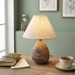May include: A ceramic table lamp with a beige linen shade sits on a wooden desk. The lamp has a teardrop-shaped base with brown and tan tones. A cup of tea, a notebook, and a notepad are also on the desk.