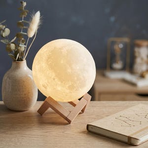 May include: A glowing moon-shaped lamp with a textured surface, resting on a wooden stand. The lamp emits a soft, warm light. A beige vase with dried plants and a notebook with constellation illustrations are also visible.