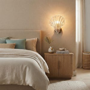 Peut inclure: Une applique murale en forme de coquillage en verre transparent, diffusant une lueur chaude. Le luminaire est placé au-dessus d'une table de chevet en bois clair avec des détails cannelés verticaux, un petit vase et des livres empilés.