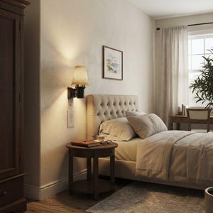 Può includere: Una camera da letto con testiera beige trapuntata, un comodino rotondo in legno scuro con un libro e occhiali, e una lampada da parete con paralume in tessuto beige. A sinistra un alto armadio in legno scuro e un quadro appeso alla parete.