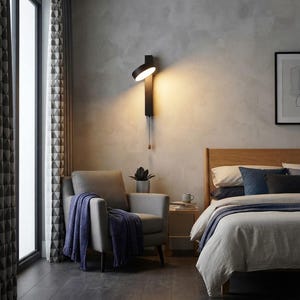 Peut inclure: Une applique murale noire avec une tête lumineuse circulaire et un interrupteur à tirette. La lampe présente une plaque de base rectangulaire verticale et émet une lueur chaude et focalisée contre le mur, au-dessus d'un fauteuil gris avec un plaid violet.