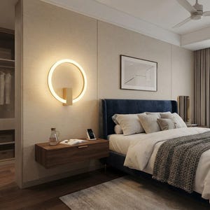 Op de afbeelding: Een cirkelvormige wandlamp met een warm oplichtend ringontwerp, voorzien van een centrale rechthoekige montagebasis in een licht houten afwerking, geplaatst boven een zwevende houten nachtkastje in een slaapkamer.
