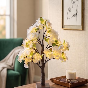 Peut inclure: Un arbre décoratif à LED avec des fleurs blanches et des feuilles vertes. Le tronc est marron et repose sur une base carrée. Les fleurs illuminées créent une lueur chaleureuse, idéale pour la décoration intérieure. L'arbre est sur une surface en bois.
