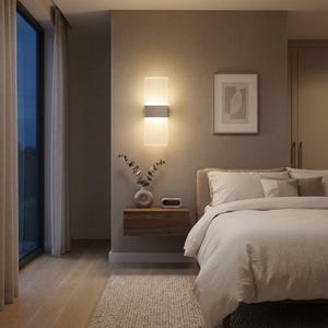 Puede incluir: Un aplique de pared rectangular con pantalla de vidrio esmerilado y una banda metálica central que emite una luz cálida. La lámpara está montada en una pared beige texturizada sobre una mesita de noche flotante de madera, junto a una cama.
