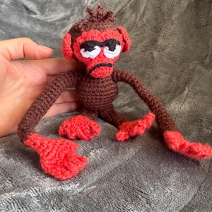 Op de afbeelding: Een handgemaakte gehaakte apenpop met een rood gezicht, handen en voeten en een bruin lichaam. De aap heeft een chagrijnige uitdrukking met zwarte ogen en een zwarte mond. De armen en benen van de pop zijn lang en slank.