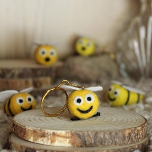 Peut inclure: Plusieurs porte-clés abeilles en feutre jaune avec des détails noirs et blancs, notamment des visages souriants et des ailes. Les porte-clés sont présentés sur de petites tranches de bois, avec un porte-clés doré attaché à l'un d'eux.