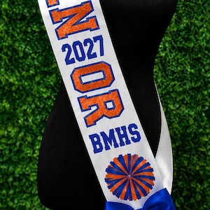 May include: A white satin sash featuring the blue text Kadie, the orange glitter text SENIOR, the blue text 2027, the blue text BMHS, a circular blue and orange pom-pom graphic, and a large blue satin bow attached at the bottom end of the sash.