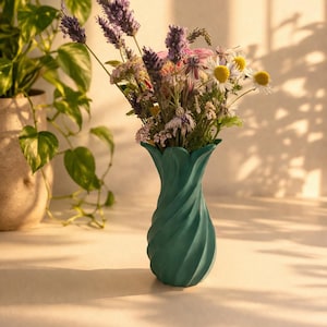 Könnte beinhalten: Eine blaugrüne Vase mit spiralförmigem, gedrehtem Design enthält einen Strauß Wildblumen, darunter violette Lavendelstängel und weiße Gänseblümchen mit gelben Mitten. Die Vase hat einen ausgestellten, blütenblattartigen Rand und eine matte Oberfläche.