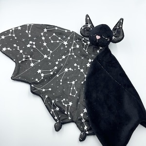 Puede incluir: Una manta para bebé con forma de murciélago. Las alas son negras con un estampado de constelaciones blancas. La cabeza del murciélago es negra con detalles rosas en la nariz y las orejas. Para bebés y niños pequeños.