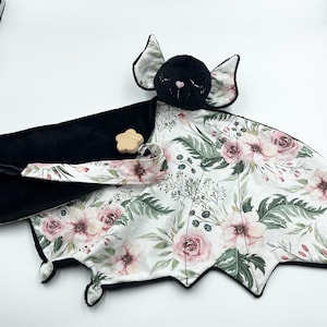 May include: A plush baby bat-shaped security blanket. The bat's head is black with pink accents, and the wings feature a floral pattern with pink roses and green leaves on a white background. A wooden star-shaped teether is attached.