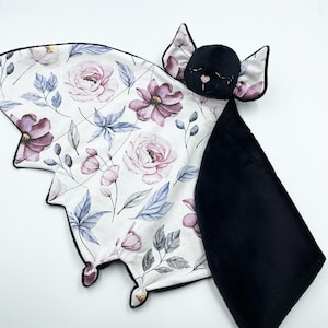 May include: A baby security blanket shaped like a bat, featuring a floral pattern in pink, purple, and blue on a white background. The bat's head is black, with small ears and a stitched face. The underside is black.