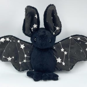 May include: A plush black bat with large ears and outstretched wings. The wings are patterned with white stars and constellations on a dark gray background. The bat has a soft, furry texture and is designed to be a cuddly toy.