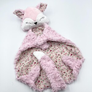 Op de afbeelding: Een roze en witte babyknuffel in de vorm van een vos. De vossenkop heeft gesloten ogen en een zwarte neus. De deken heeft een bloemenpatroon aan de binnenkant en een getextureerd, roosachtig patroon aan de buitenkant. De staart is ook van pluche.