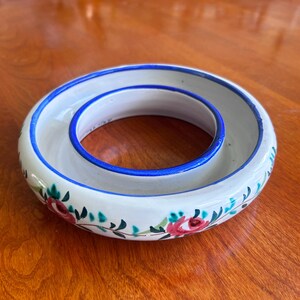 May include: A circular ceramic ring-shaped dish with a white glazed finish, featuring hand-painted red rose motifs and green leaves around the exterior. The inner and outer rims are accented with a thin, vibrant blue painted border line.