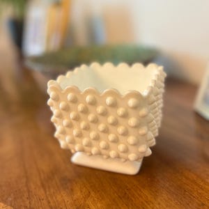 May include: A square-shaped off-white milk glass planter featuring a raised hobnail texture on all four sides and a scalloped top edge. The vessel sits on a small, solid square base and has a smooth interior finish.