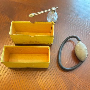 May include: A clear glass perfume atomizer with a silver-toned metal nozzle, a gray rubber bulb with a black hose, and a two-piece rectangular yellow cardboard box. The components are separate, showing the glass bottle, the pump, and the box.