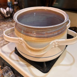 May include: A ceramic cooking pot with a wide opening and two side handles. The pot features a cream-colored base with a blue horizontal line, transitioning to a tan and brown glazed rim. The handles have a decorative four-dot embossed pattern.