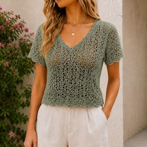 May include: A sage green crochet top with a V-neck and short sleeves. The top has a scalloped hemline and a delicate, openwork design. The model is wearing the top with beige pants.