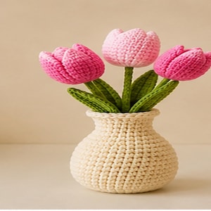 Op de afbeelding: Een gehaakte vaas bevat drie handgemaakte tulpenbloemen. De tulpen zijn roze, met groene stelen en bladeren. De vaas is gebroken wit en heeft een getextureerd, geweven uiterlijk. De achtergrond is effen neutraal.
