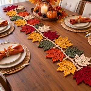 May include: A crochet table runner featuring a repeating pattern of maple leaves in shades of red, orange, yellow, green, and cream. The runner is laid across a wooden dining table set with plates, orange napkins, and a centerpiece with a candle.