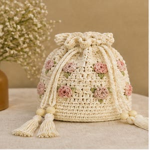 May include: Cream-colored crochet drawstring bag adorned with pink rose appliqués and green leaves. The bag features a drawstring closure and cream-colored tassels with wooden beads. The bag is sitting on a neutral surface.