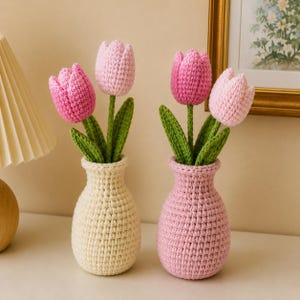 Puede incluir: Dos jarrones de ganchillo, uno crema y otro rosa, cada uno con dos o tres tulipanes de ganchillo. Los tulipanes tienen pétalos rosas y tallos y hojas verdes. Los jarrones están sobre una superficie blanca.